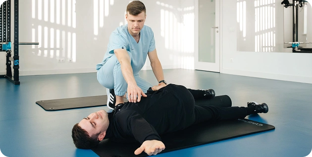 De revalidatietherapeut helpt de man bij het uitvoeren van rekoefeningen en oefeningen met een massageroller (foamroller) op een matje.